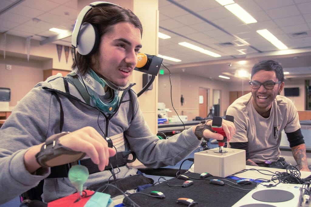 Picture of a disabled indidivual using adaptive equipment, and a man sat next to him helping him out. This is to support the inclusion of disabled people in esports